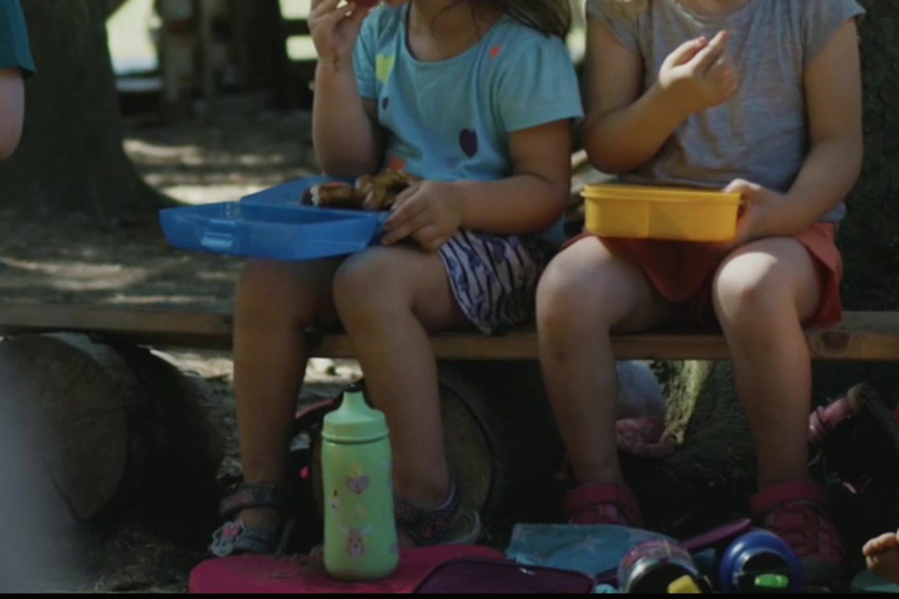 Kinder sitzen mit ihren Brotzeitdosen auf der Bank am Waldplatz